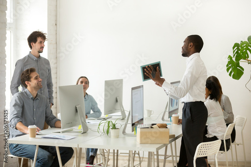 New african employee unpacking boxes and talking to friendly colleagues on first working day in office, smiling happy black worker having pleasant conversation with coworkers showing photo in frame