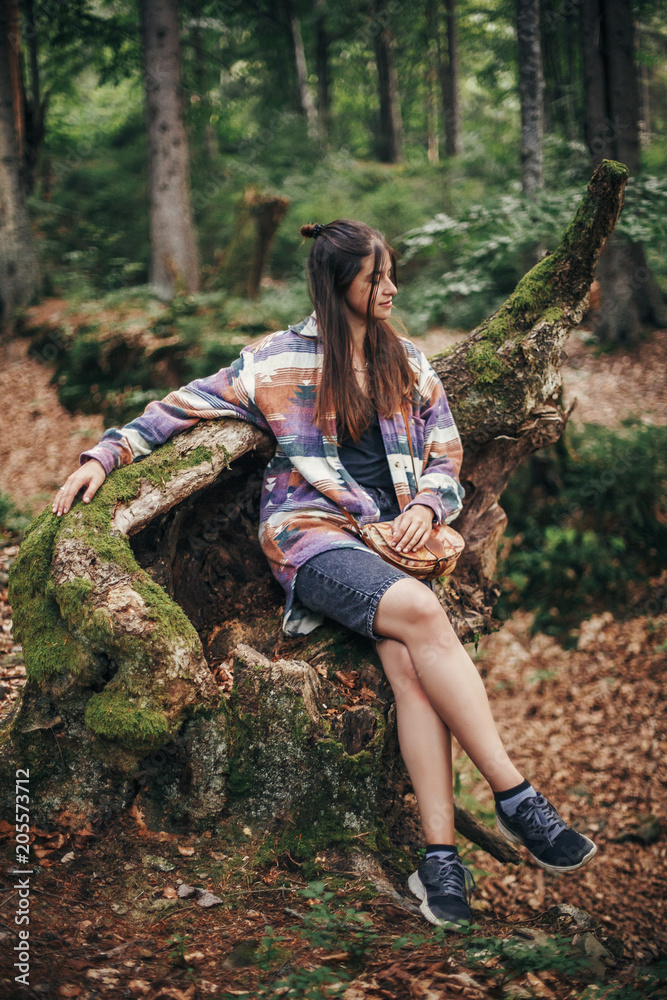 stylish traveler girl sitting on old stump with moss in sunny forest and mountains. travel and wanderlust concept. space for text. happy hipster woman relaxing in woods stylish traveler girl sitting on old stump with moss in sunny forest and mountains. travel and wanderlust concept. space for text. happy hipster woman relaxing in woods