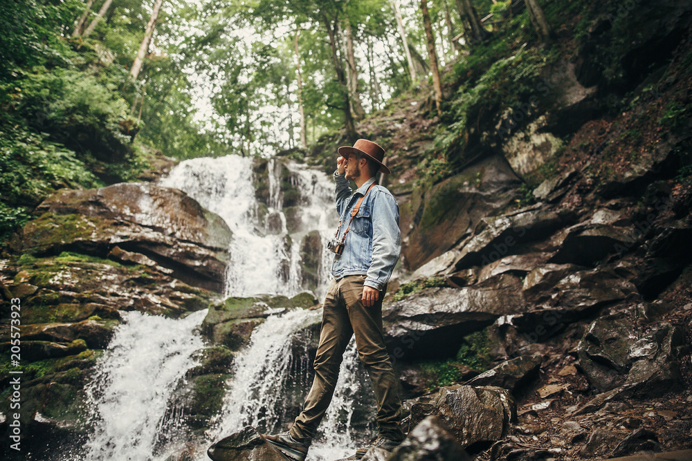 travel and wanderlust concept. stylish hipster man in hat with photo camera, standing at waterfall in forest in mountains. traveler guy exploring woods. space for text