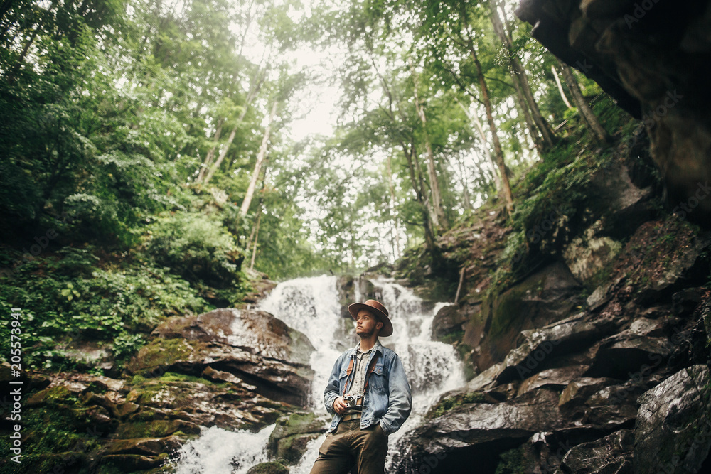 travel and wanderlust concept. stylish hipster man in hat with photo camera, standing at waterfall in forest in mountains. traveler guy exploring woods. space for text