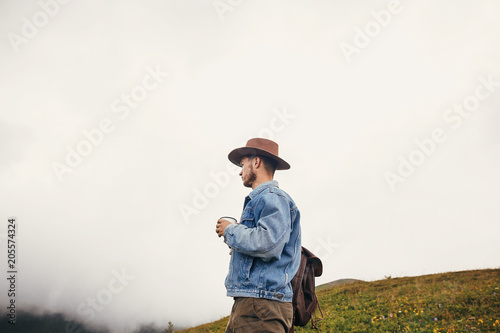 travel and wanderlust concept. stylish traveler man in hat with backpack holding cup of tea in mountains with clouds. space for text. hipster guy traveling. amazing atmospheric moment