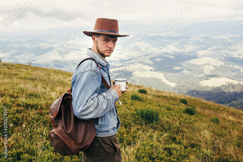 stylish traveler man in hat with backpack holding cup of tea in mountains with clouds. space for text. hipster guy traveling. amazing atmospheric moment. travel and wanderlust concept