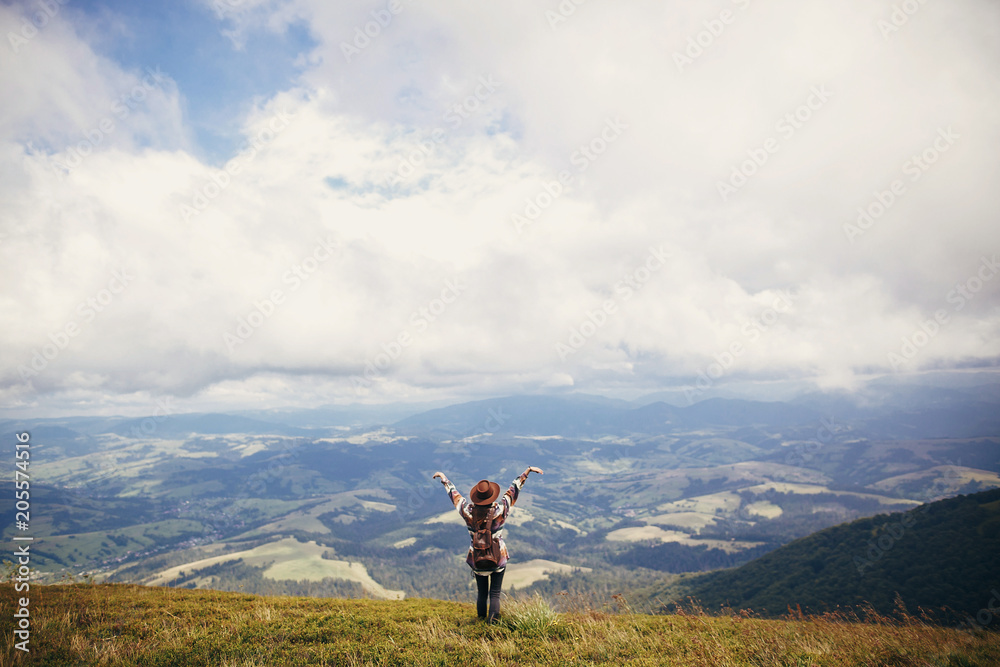 happy traveler hipster girl in hat, standing with backpack and raising hands up in sky in mountains in clouds. space for text. amazing atmospheric emotional moment. travel and wanderlust concept