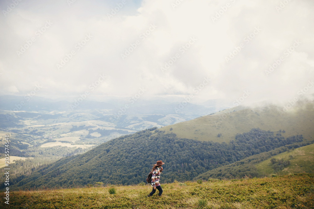 happy stylish traveler girl holding hat with backpack and windy hair in mountains clouds. summer vacation. travel and wanderlust concept. space for text. amazing atmospheric moment happy stylish traveler girl holding hat with backpack and windy hair in mountains clouds. summer vacation. travel and wanderlust concept. space for text. amazing atmospheric moment