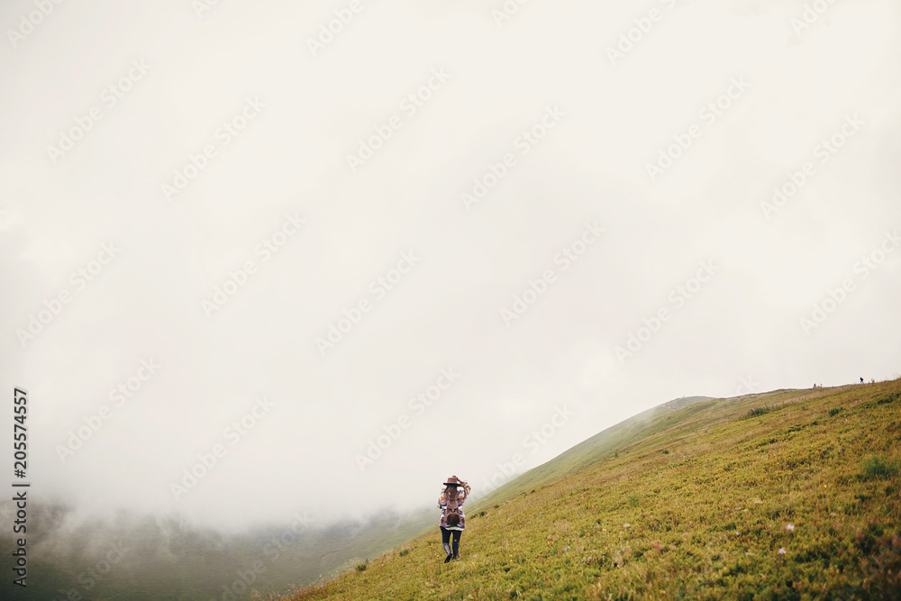 stylish traveler girl holding hat with backpack and windy hair, walking in mountains clouds. summer vacation. travel and wanderlust concept. space for text. amazing atmospheric moment