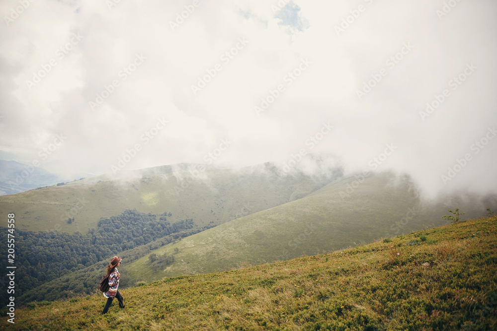 happy traveler hipster girl in hat, walking with backpack in sunny mountains in the  clouds. space for text. amazing atmospheric emotional moment. travel and wanderlust concept
