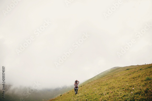 Fototapeta Naklejka Na Ścianę i Meble -  stylish traveler girl holding hat with backpack and windy hair, walking in mountains clouds. summer vacation. travel and wanderlust concept. space for text. amazing atmospheric moment