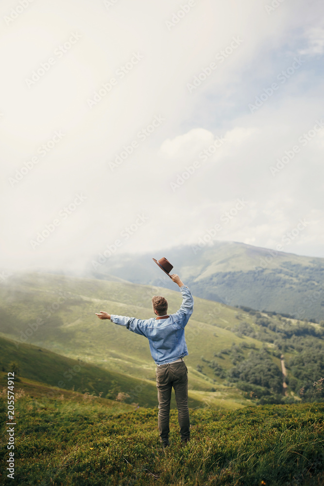 travel and wanderlust concept. stylish traveler man holding hat standing on top of sunny mountains in clouds. space for text. hipster guy traveling. amazing atmospheric moment.