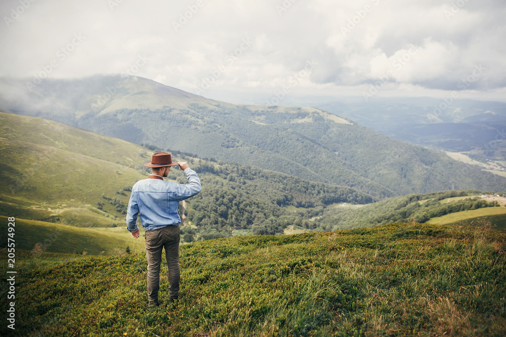 stylish traveler man in hat standing on top of sunny mountains in clouds. space for text. hipster guy traveling. amazing atmospheric moment. travel and wanderlust concept