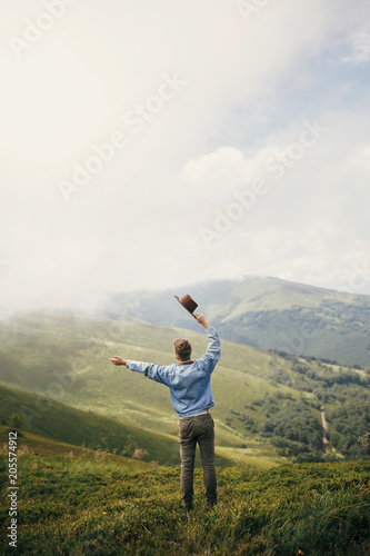travel and wanderlust concept. stylish traveler man holding hat standing on top of sunny mountains in clouds. space for text. hipster guy traveling. amazing atmospheric moment.