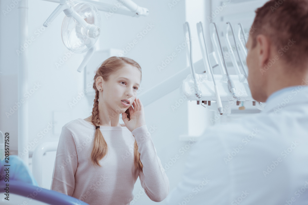 Dull pain. Melancholy dreary girl touching face while gazing at male dentist Dull pain. Melancholy dreary girl touching face while gazing at male dentist