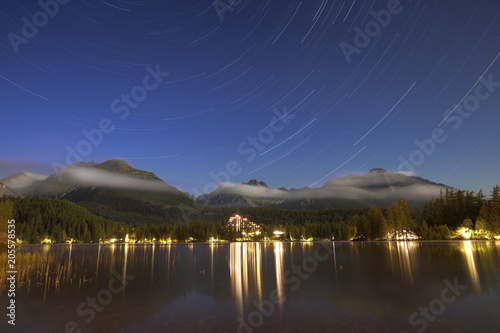 Majestic view of lake by illuminated town against mountains and star trails at night photo