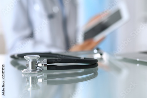 Stethoscope lying on glass desk with laptop computer at busy physician background. Medicine or pharmacy concept. Medical tools at doctor working table