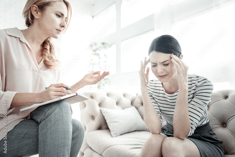 Big problem. Unhappy pretty exhausted woman sitting on the sofa near another woman touching to her head and feeling bad herself.