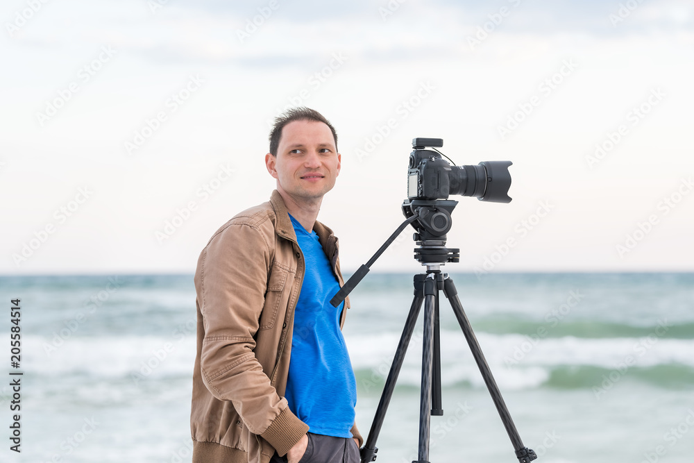 Young man professional photographer taking pictures and video of beach sunset in Florida panhandle, with jacket, wind, beach waves, tripod