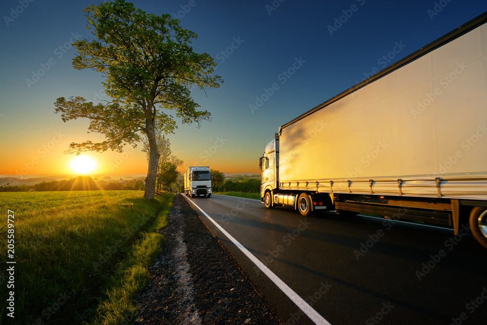 Trucks driving on the asphalt road between trees in a rural landscape at sunset