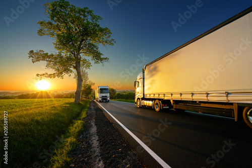Trucks driving on the asphalt road between trees in a rural landscape at sunset