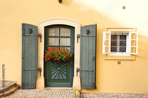 A yellow house wall with blue wood door and window on a small street. On the door hangs a flower box with red flowers