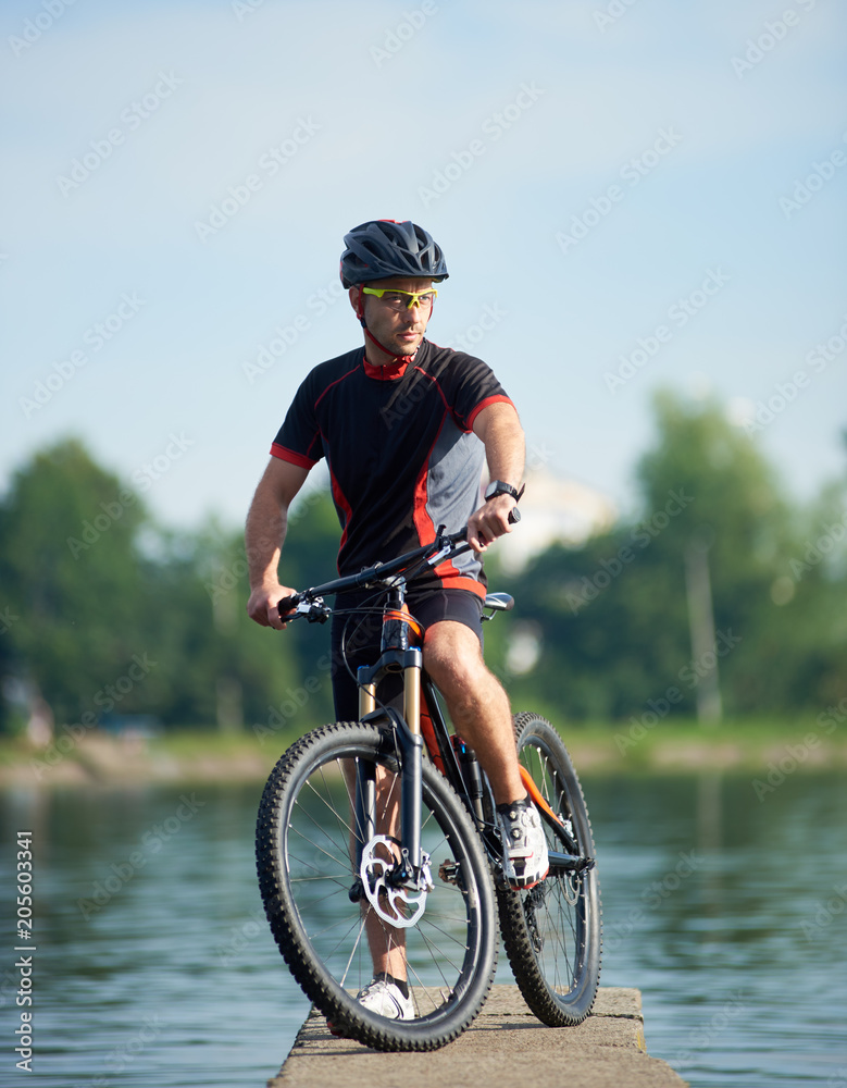 Male bicyclist in professional cycling sportswear and protective helmet posing near bicycle looking away, thinking about future success and achievement. Sportsman taking break at pier near lake.