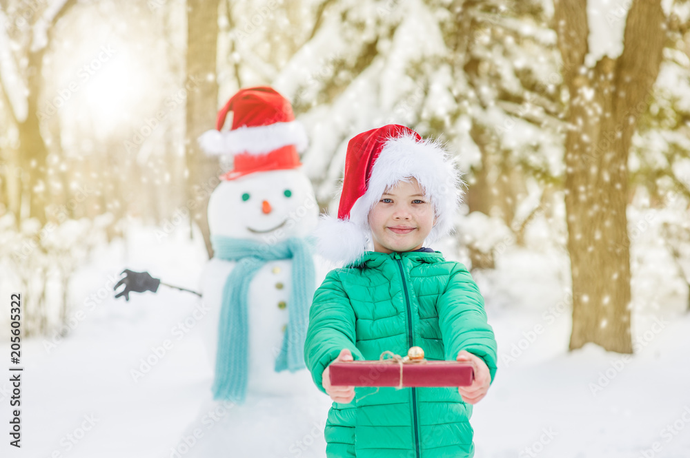 portrait of smiling boy in red christmas hat holds gift box with snowman on background