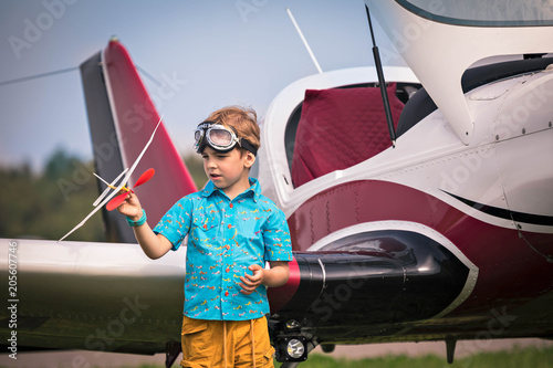 Caucasian boy  in yellow shorts, a blue shirt with the drawn small planes and in aviation points holds the toy plane in hand and dreming. On a background the real plane of small aircraft. photo