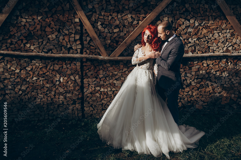 Fototapeta premium rustic wedding couple embracing in sunlight at background of wooden firewood. barn wedding concept, space for text. happy stylish bride and groom at wood wall, romantic sensual moment