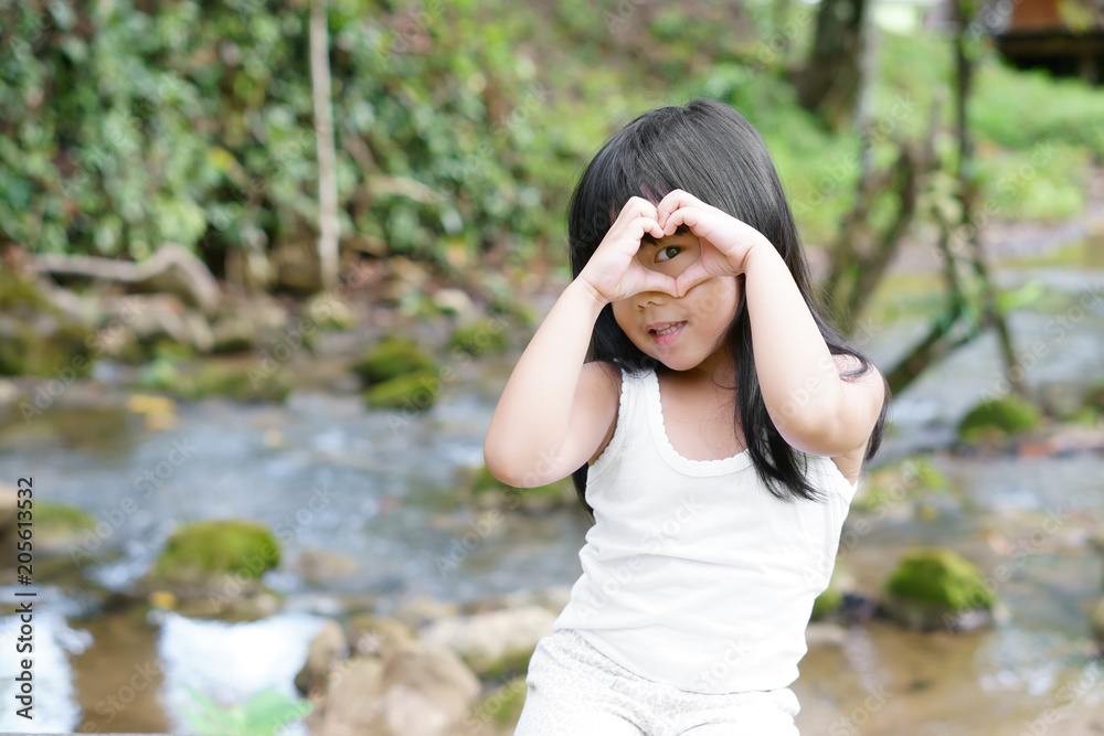 Asian children cute or kid girl smile with wear white vest and make hand heart on eye for love on stream nature in garden or green jungle with holiday relax on summer travel trip Asian children cute or kid girl smile with wear white vest and make hand heart on eye for love on stream nature in garden or green jungle with holiday relax on summer travel trip