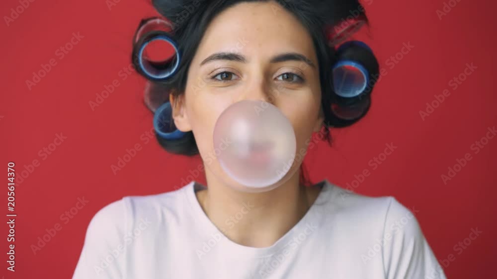 Girl with curlers on her head and gum blowing bubble on red background.