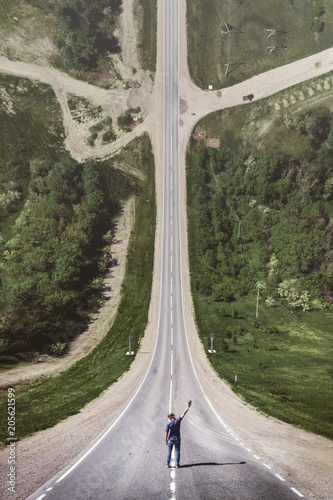 A young man in a cap with a hand raised up with a console from a drone is standing on a countryside asphalt road at the end of which there is a crossroads. Photo in the style of the film Inception