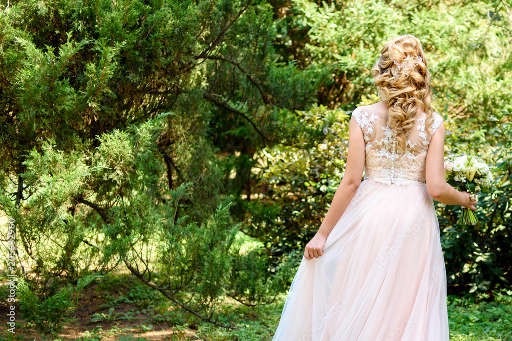 Beautiful bride in wedding dress with bridal bouquet in the park outdoors,  back view. Blond girl with curly hair styling and jewelry. Wedding hairstyle  for long hair with stylish hair accessory Stock