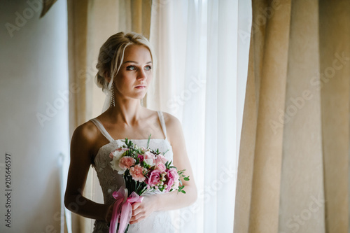 Serious portrait of the beautiful bride with a wedding bouquet of flowers near the window indoors in the rooms.