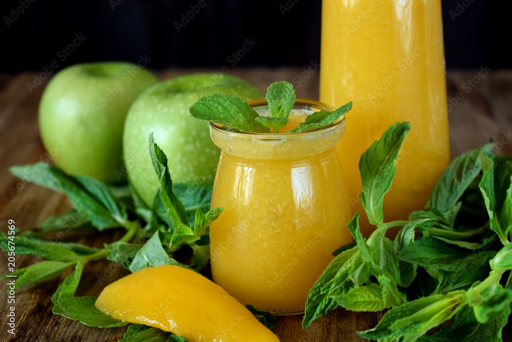 Fototapeta premium Yellow smoothie in glass jars surrounded by fruits and mint against wooden background