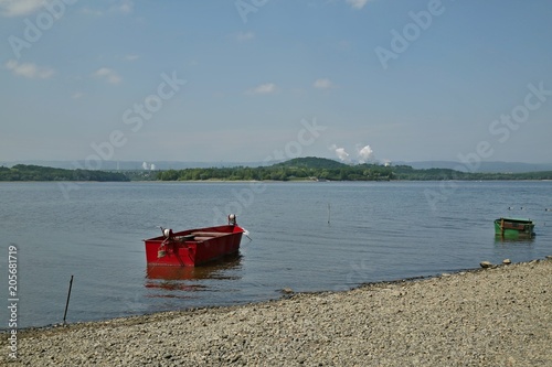 Landscape with Nechranice dam, Czech Republic, sandy coastline with pebbles, red, green fisherman boat on blue water with waves, horizon with green trees, white smoke and cooling towers of power plant