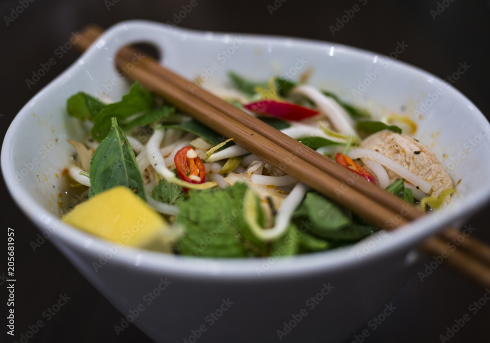 Beautiful and appetizing photo of a traditional vietnamese chicken noodle soup, also know as Pho Ga. In a white bowl with wooden background and luxurious wooden chopsticks.