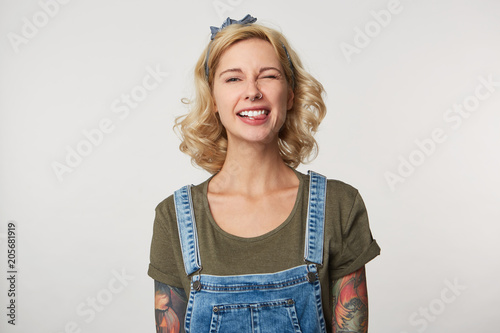 Cute blonde female  wears denim overalls, casual t shirt, smiles and shows her tongue, feels happy, funny photo