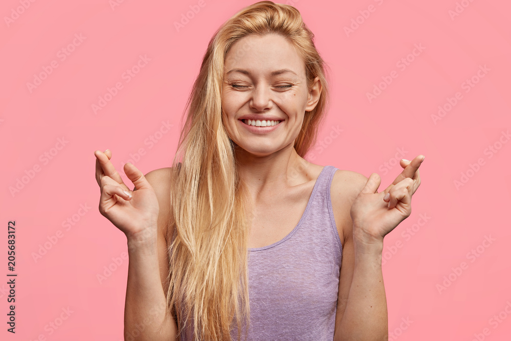Indoor portrait of hopeful glad female with long light hair, keeps fingers crossed, hopes for good luck before important event, has broad smile, isolated over pink background. Body language concept Indoor portrait of hopeful glad female with long light hair, keeps fingers crossed, hopes for good luck before important event, has broad smile, isolated over pink background. Body language concept