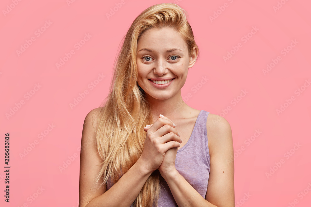 Positive young blonde woman keeps hands together, smiles gladfully, happy to recieve good news from someone, dressed casually, demonstrates her beauty and pleasant emotions, isolated on pink Positive young blonde woman keeps hands together, smiles gladfully, happy to recieve good news from someone, dressed casually, demonstrates her beauty and pleasant emotions, isolated on pink