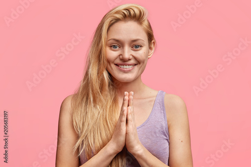 Horizontal shot of happy young European woman with no make up, keeps hands pressed together, makes praying gesture as asks for forgiveness, has cheerful expression, isolated on pink background