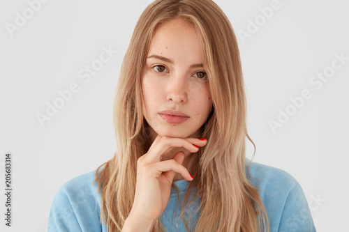 Portrait of lovely young female with serious expression, holds chin, looks confidently at camera, poses against white background, thinks about something. Pretty woman designer has new idea for project