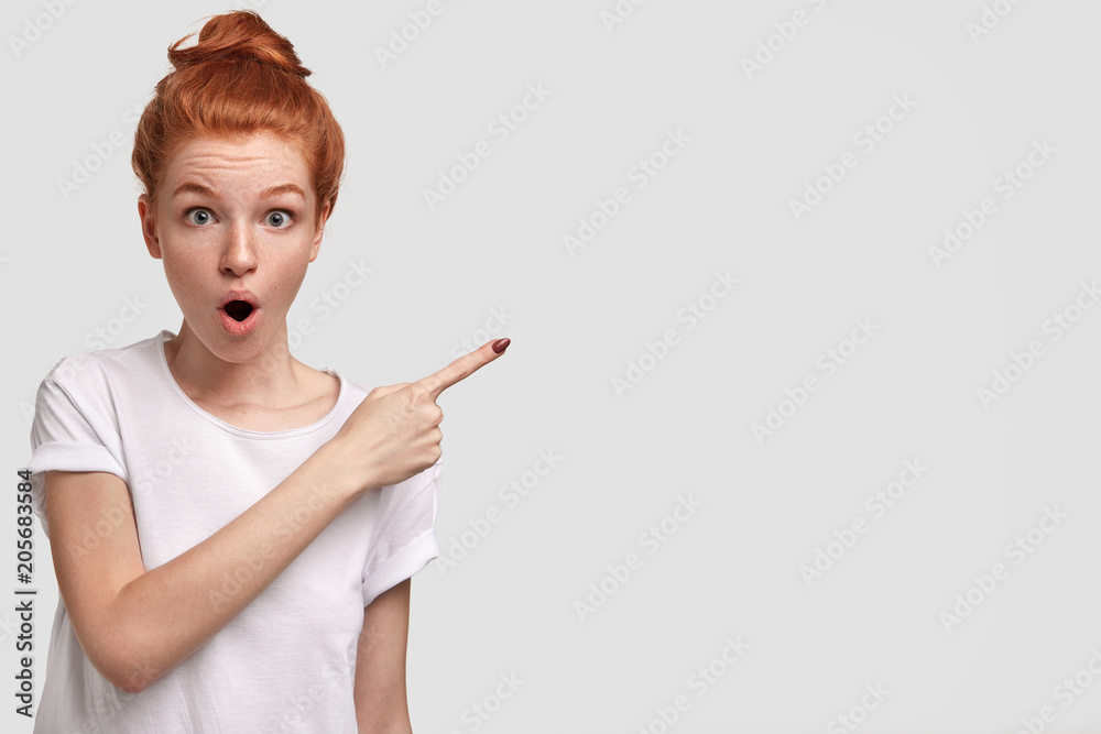 Studio shot of attractive young freckled female with red hair combed in knot, indicates on right at blank copy space, has stunned expression and jaw dropped, sees something amazing or wonderful Studio shot of attractive young freckled female with red hair combed in knot, indicates on right at blank copy space, has stunned expression and jaw dropped, sees something amazing or wonderful
