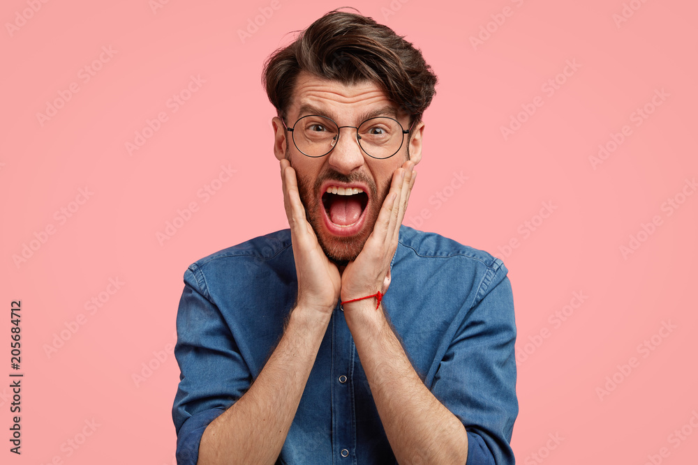 Portrait of outraged furious aggressive man screams angrily, has annoyed expression, keeps mouth widely opened, being crazy and mad, wears fashionable clothes, stands against pink background. Portrait of outraged furious aggressive man screams angrily, has annoyed expression, keeps mouth widely opened, being crazy and mad, wears fashionable clothes, stands against pink background.