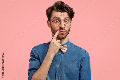 Attractive bearded male looks with eyes full of disbelief, keeps fore finger near mouth, has attractive appearance, wears formal clothes, stands against pink background, excited with bad news