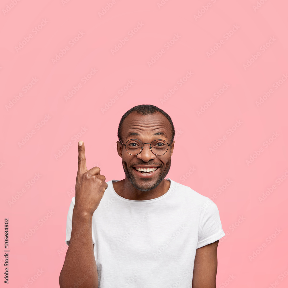 Look upwards! Satisfied glad middle aged African American male points with fore finger upwards, being excited as sees and advertises something awesome, isolated on pink background with copy space Look upwards! Satisfied glad middle aged African American male points with fore finger upwards, being excited as sees and advertises something awesome, isolated on pink background with copy space