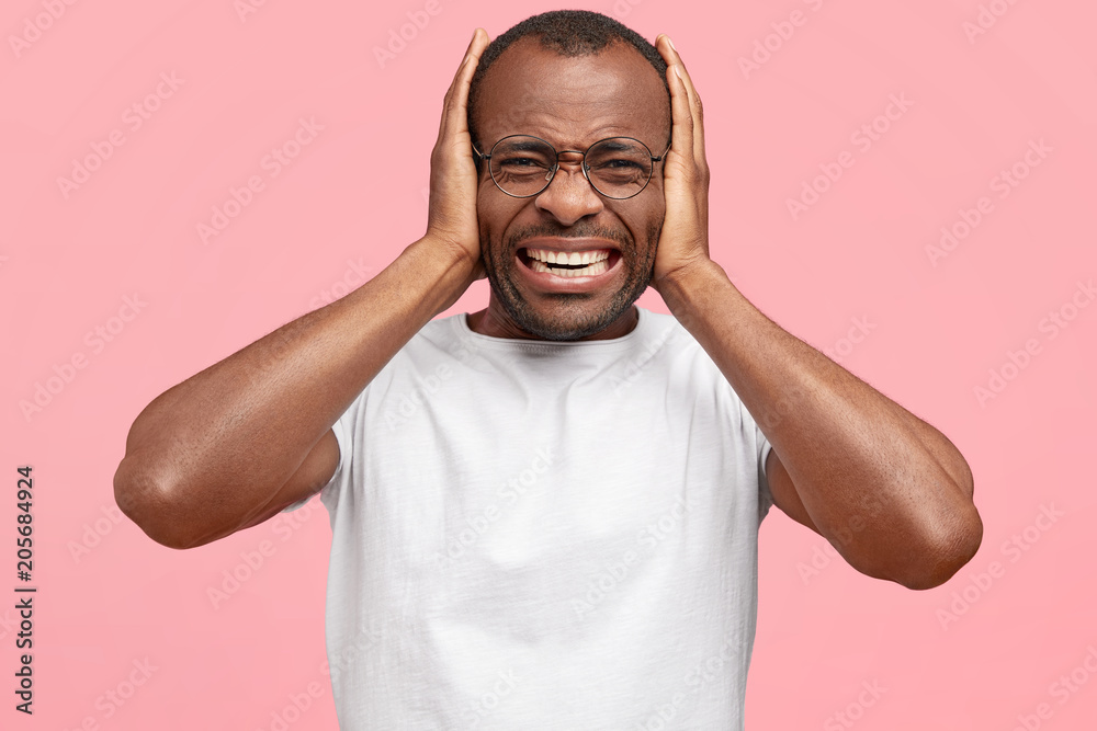 Stressed desperate dark skinned male with sorrowful annoyed expression, covers ears, being in panic as hears terrified news, wears white t shirt, isolated on pink background. Negative human emotions Stressed desperate dark skinned male with sorrowful annoyed expression, covers ears, being in panic as hears terrified news, wears white t shirt, isolated on pink background. Negative human emotions
