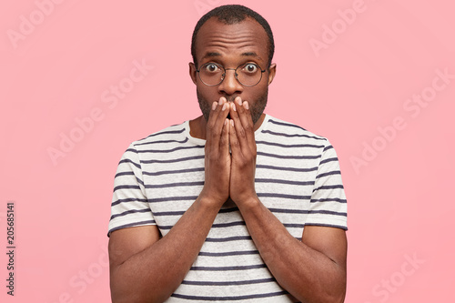 Tableau sur toile Frightened African American male looks with scared expression, covers mouth, wears casual clothes, being afraid of something, poses against pink background