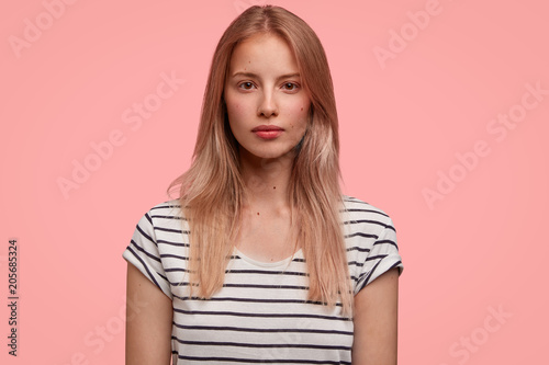 Pretty young female model with satisfied expression, wears casual t shirt, being serious, isolated over pink background. Attractive European student has fair hair, listens attentively lecture