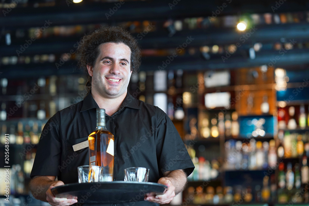 A bartender man carries a bottle of whiskey on a tray to the client in the hotel bar. The concept of service.
