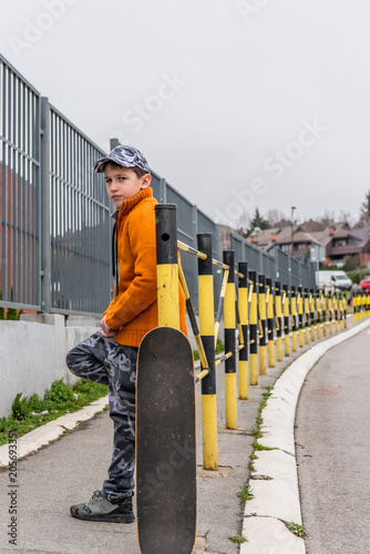 Young boy with his skateboard waiting, leaning on street fence