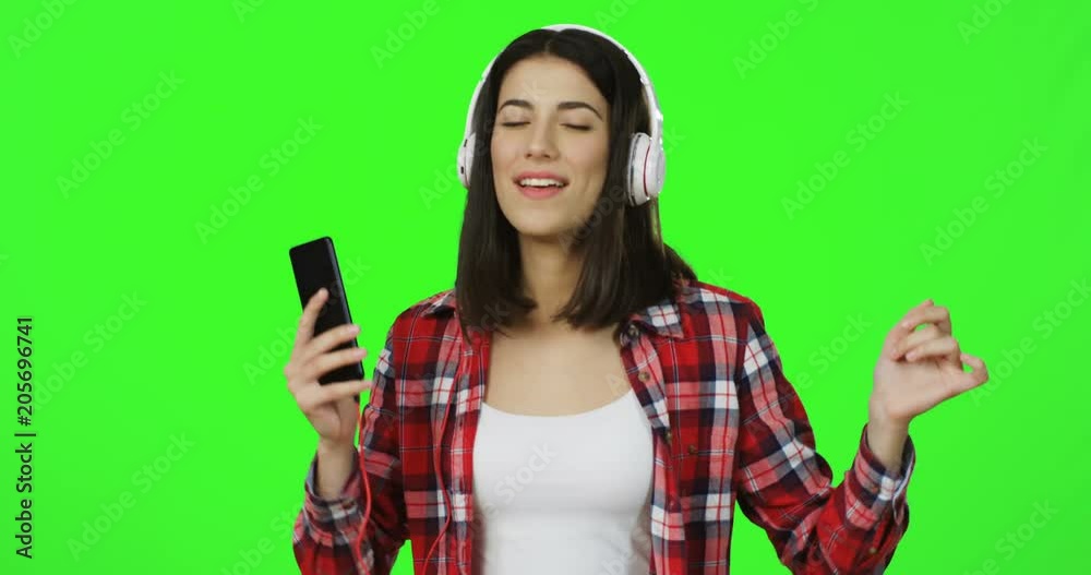 Portrait shot of the brunette attractive young woman in the red plaid shirt and hedphones listening to the music on the smartphone and dancing while standing in front of the camera on the green screen Portrait shot of the brunette attractive young woman in the red plaid shirt and hedphones listening to the music on the smartphone and dancing while standing in front of the camera on the green screen