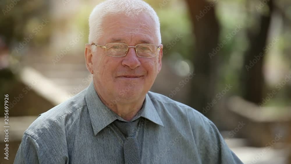 Portrait of a happy aged man in a grey shirt and glasses smiling, standing, and feeling good in a green street on a sunny day in summer Portrait of a happy aged man in a grey shirt and glasses smiling, standing, and feeling good in a green street on a sunny day in summer